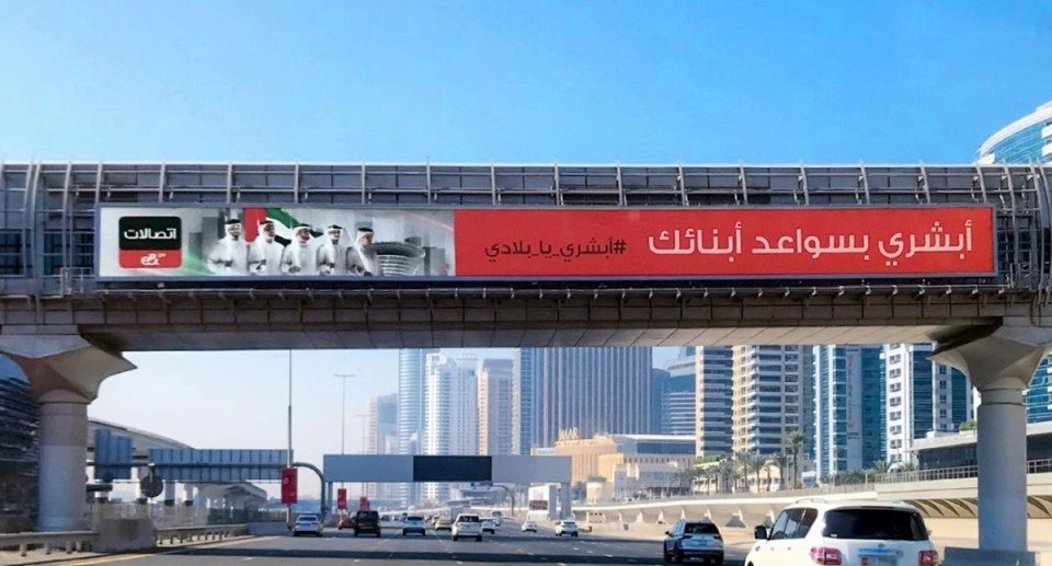 Long horizontal billboard mounted on road bridge with Arabic advertising copy above busy urban roadway