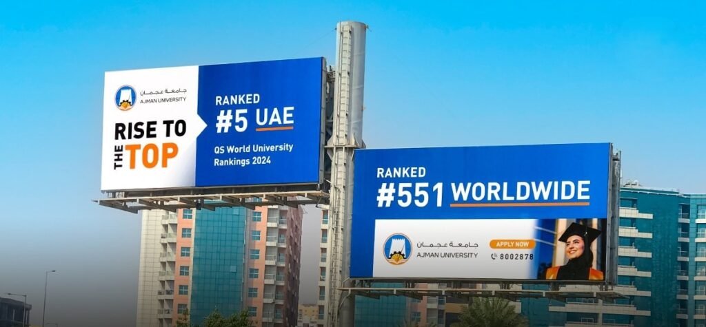 Twin roadside billboards promoting Ajman University rankings positioned near urban buildings and traffic routes.