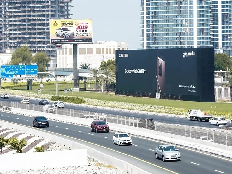 Illuminated roadside billboard displaying public messaging placed along an Ajman street with surrounding traffic.