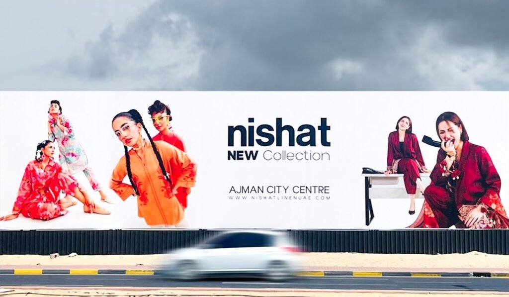 Outdoor advertising billboard installed beside a wide Ajman road with commercial buildings and passing vehicles.