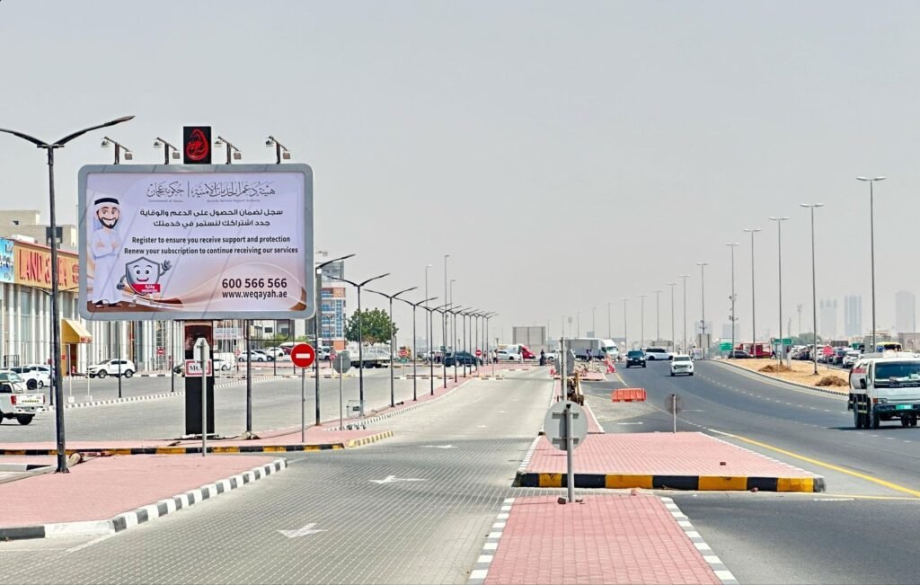 Street-level government awareness billboard placed near a major Ajman roadway with vehicles and pedestrian paths.