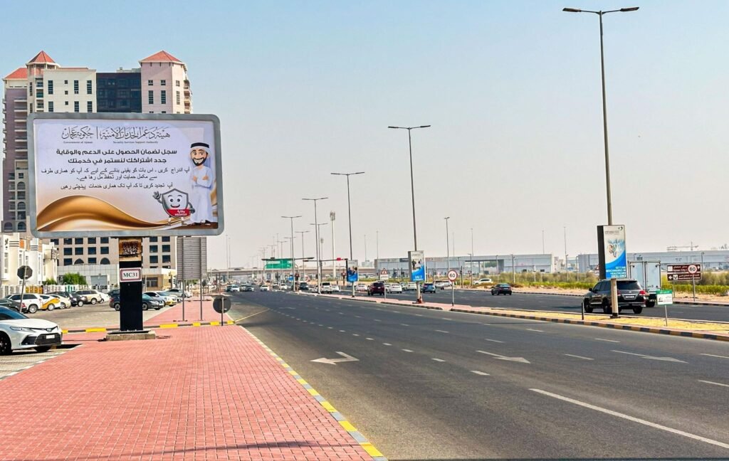 Street advertising billboard positioned along a multi-lane Ajman road with visible traffic flow and sidewalks.